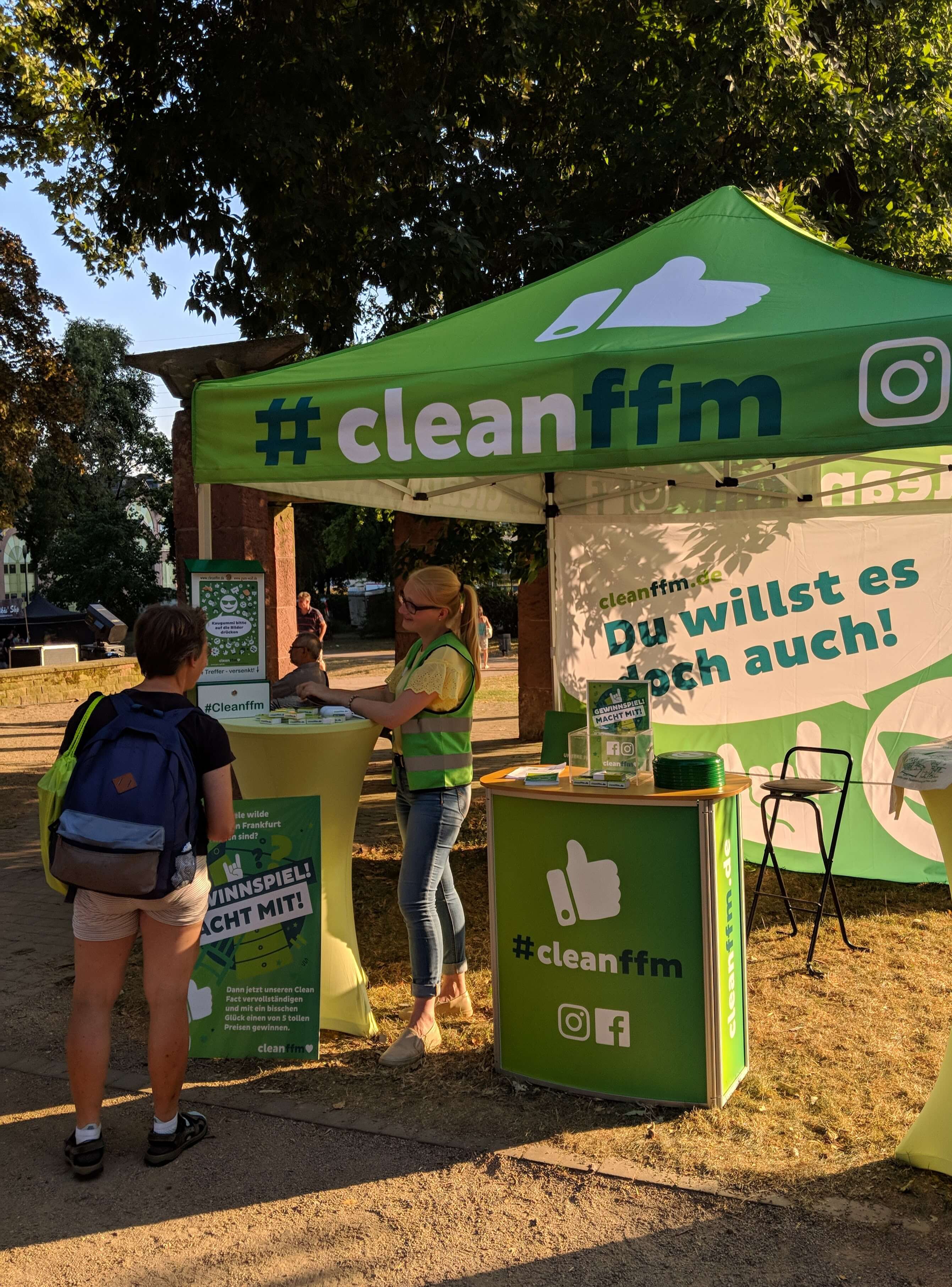 Infostand unter grünem Pavillon, Sauberkeitsbotschafterin spricht mit Besuchenden