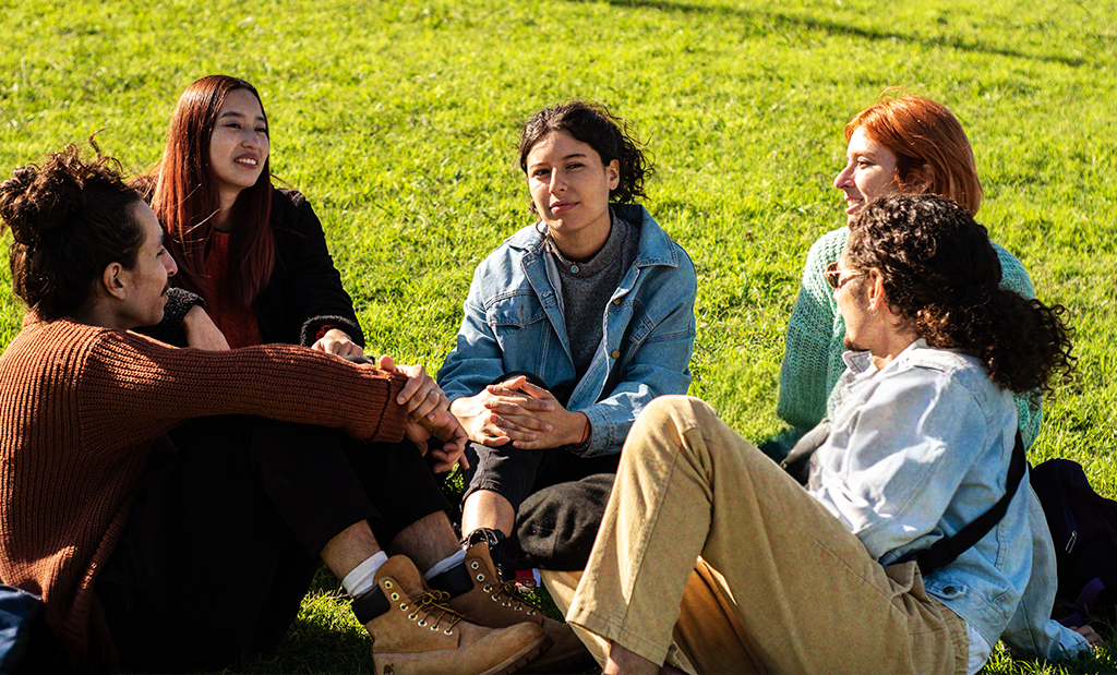 Foto: Fünf junge Menschen sitzen lachend im Gras und unterhalten sich entspannt in der Sonne.